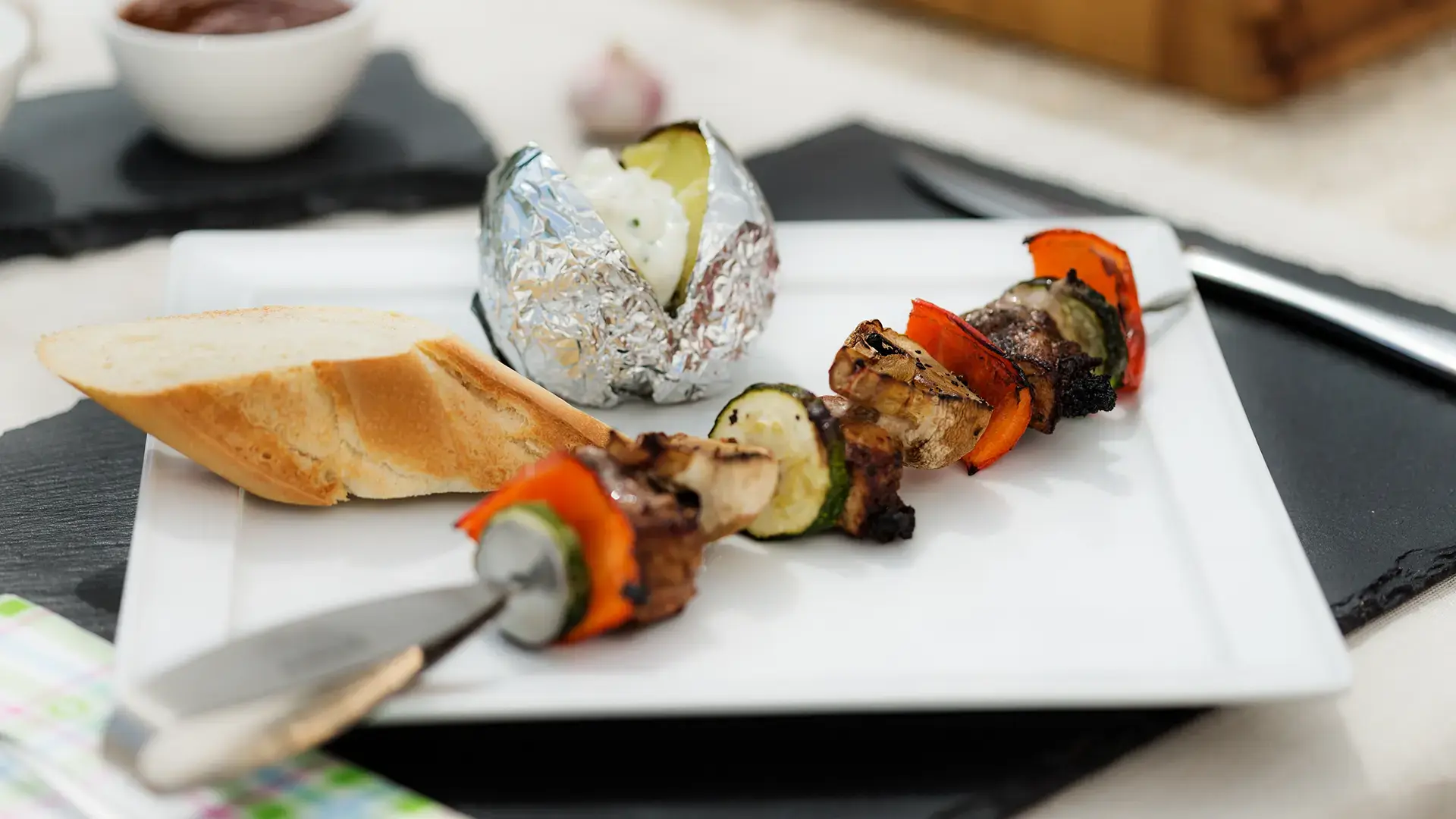 Rottaler Grillspieß mit Strohsau und Weiderind auf Edelstahlspieß, serviert mit Folienkartoffel und Rottaler Weißbrot auf Schiefer